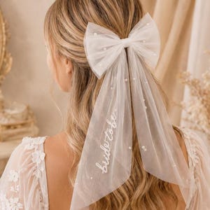 May include: A white tulle hair bow featuring scattered faux pearls and two long, flowing ribbons. One ribbon is embroidered with the white cursive text bride to be. The accessory is attached to a hair clip and is worn in long, wavy blonde hair.