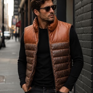 May include: A sleeveless puffer gilet featuring a two-tone design with a smooth tan leather upper section and a quilted dark brown leather lower section. The gilet has a high collar, a full-length front zip, and a structured, padded construction.