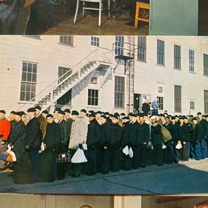 May include: A black and white photograph of a group of men in uniform standing in a line in front of a building. The men are wearing black hats and dark jackets. The building is a large, white building with many windows.