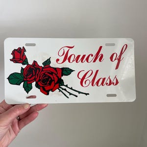 May include: White license plate with red roses and the text "Touch of Class" in red script. The roses have green leaves and stems. The plate has four mounting holes.