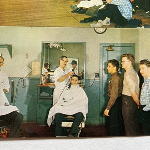 May include: A barber shop scene with a barber giving a haircut to a man sitting in a chair. There are other men waiting for their turn. The barber is using an electric razor. The walls are painted a light blue color.