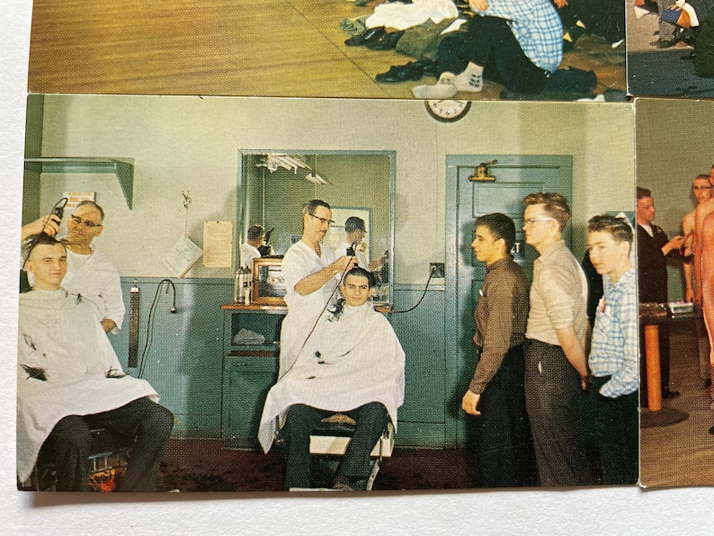 May include: A barber shop scene with a barber giving a haircut to a man sitting in a chair. There are other men waiting for their turn. The barber is using an electric razor. The walls are painted a light blue color.