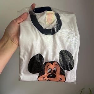 Op de afbeelding: Een wit ringer-t-shirt met een donkerblauwe kraag en een grafische print van het gezicht van Mickey Mouse op de voorzijde. Het shirt zit in een doorzichtige plastic zak en heeft een zichtbaar label bij de halslijn met de naam en maat.