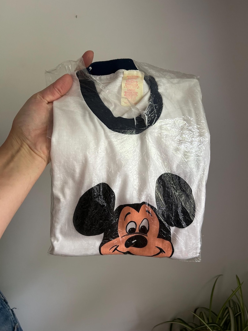 Op de afbeelding: Een wit ringer-t-shirt met een donkerblauwe kraag en een grafische print van het gezicht van Mickey Mouse op de voorzijde. Het shirt zit in een doorzichtige plastic zak en heeft een zichtbaar label bij de halslijn met de naam en maat.