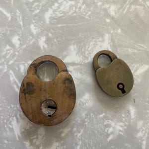 May include: Two antique brass padlocks. The larger lock has a circular body with a keyhole and a curved handle. The smaller lock has a similar design. Both locks show signs of age and wear, with a tarnished finish.