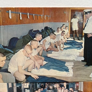 May include: A group of men in white undershirts are sitting on the floor in a room. They are inspecting their folded blue pants. A man in a white uniform is standing in the background, watching them.