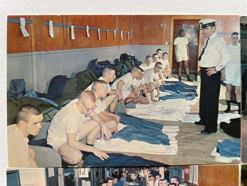 May include: A group of men in white undershirts are sitting on the floor in a room. They are inspecting their folded blue pants. A man in a white uniform is standing in the background, watching them.