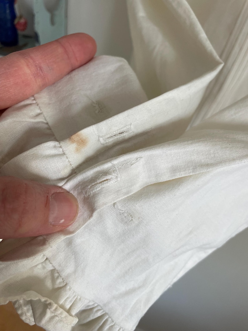 Puede incluir: Primer plano de una camisa blanca de algod&oacute;n con un ribete de volantes y dos cierres de bot&oacute;n. Hay una peque&ntilde;a mancha marr&oacute;n en la tela.