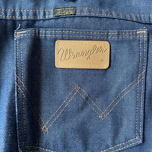 May include: Close-up of the back pocket of blue denim jeans. The pocket features a brown leather patch with the brand name "Wrangler" embossed in a cursive font. The pocket is outlined with tan stitching.