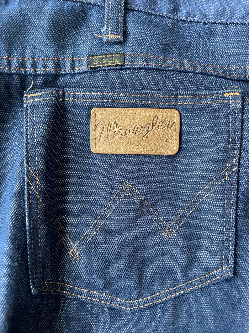 May include: Close-up of the back pocket of blue denim jeans. The pocket features a brown leather patch with the brand name "Wrangler" embossed in a cursive font. The pocket is outlined with tan stitching.