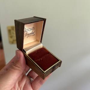 May include: A small, rectangular, dark brown jewelry box with a hinged lid. The interior features a deep red velvet ring insert and a cream-colored satin lining on the lid, which is printed with the text Lawson's Jewelers Since 1900.