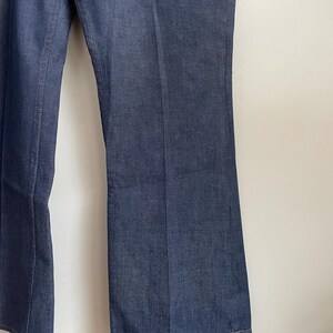 May include: Close-up of dark blue denim pants. The pants have a slight flare at the bottom and a classic five-pocket design. The denim fabric has a subtle texture, and the stitching is visible along the seams.