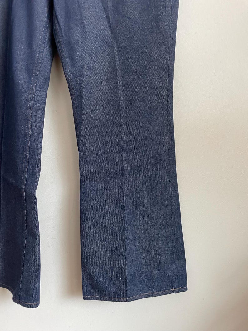 May include: Close-up of dark blue denim pants. The pants have a slight flare at the bottom and a classic five-pocket design. The denim fabric has a subtle texture, and the stitching is visible along the seams.