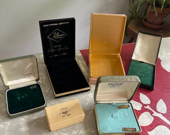 May include: A collection of six vintage jewelry boxes in various colors and sizes. The boxes feature brand names including Ballou, Jo-Anna Jewels, Lurias, and Van Dell. Some boxes are open, revealing velvet-lined interiors in green, black, and blue.