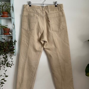 May include: A pair of beige straight-leg jeans with a classic five-pocket design. The jeans are hanging on a hanger and have a light brown color.