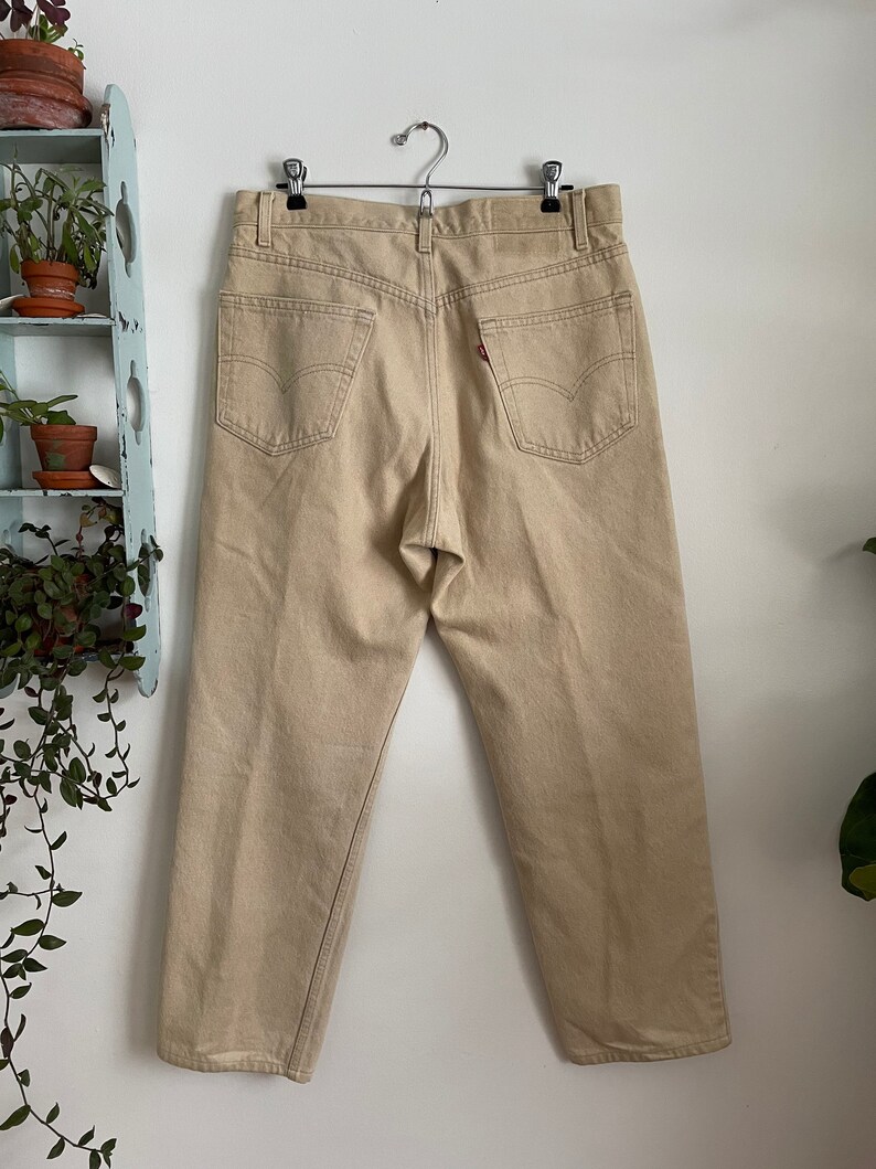 May include: A pair of beige straight-leg jeans with a classic five-pocket design. The jeans are hanging on a hanger and have a light brown color.