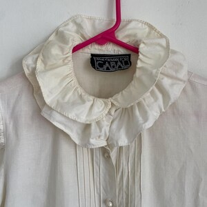 Puede incluir: Una camisa blanca de botones con un cuello grande y con volantes. La camisa tiene una etiqueta que dice "Diane Gilman for Cabal".