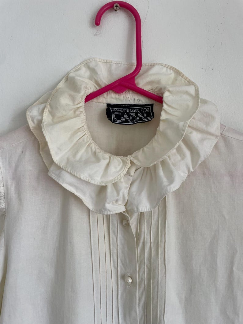 Puede incluir: Una camisa blanca de botones con un cuello grande y con volantes. La camisa tiene una etiqueta que dice "Diane Gilman for Cabal".