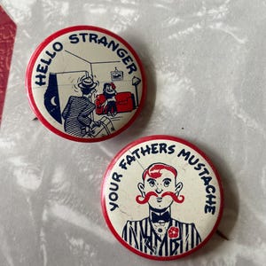 May include: Two vintage button pins. The first pin is red and white with the text "Hello Stranger" in blue. The pin features a cartoon image of a man entering a room with a woman sitting on a couch. The second pin is red and white with the text "Your Fathers Mustache" in blue. The pin features a cartoon image of a man with a large red mustache.