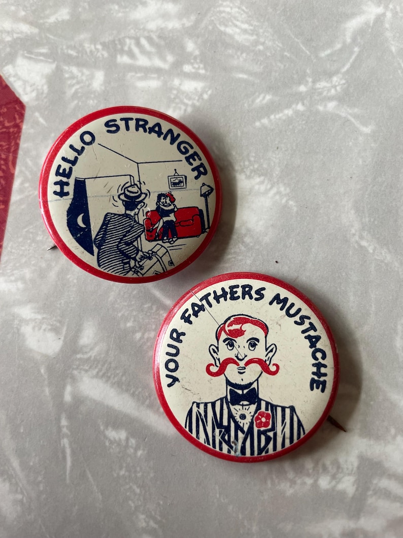 May include: Two vintage button pins. The first pin is red and white with the text "Hello Stranger" in blue. The pin features a cartoon image of a man entering a room with a woman sitting on a couch. The second pin is red and white with the text "Your Fathers Mustache" in blue. The pin features a cartoon image of a man with a large red mustache.