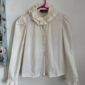 Puede incluir: Una blusa blanca de manga larga con cuello y pu&ntilde;os con volantes. La blusa tiene un cierre de botones en la parte delantera y est&aacute; plisada en la parte delantera. La etiqueta dice "CARAD"