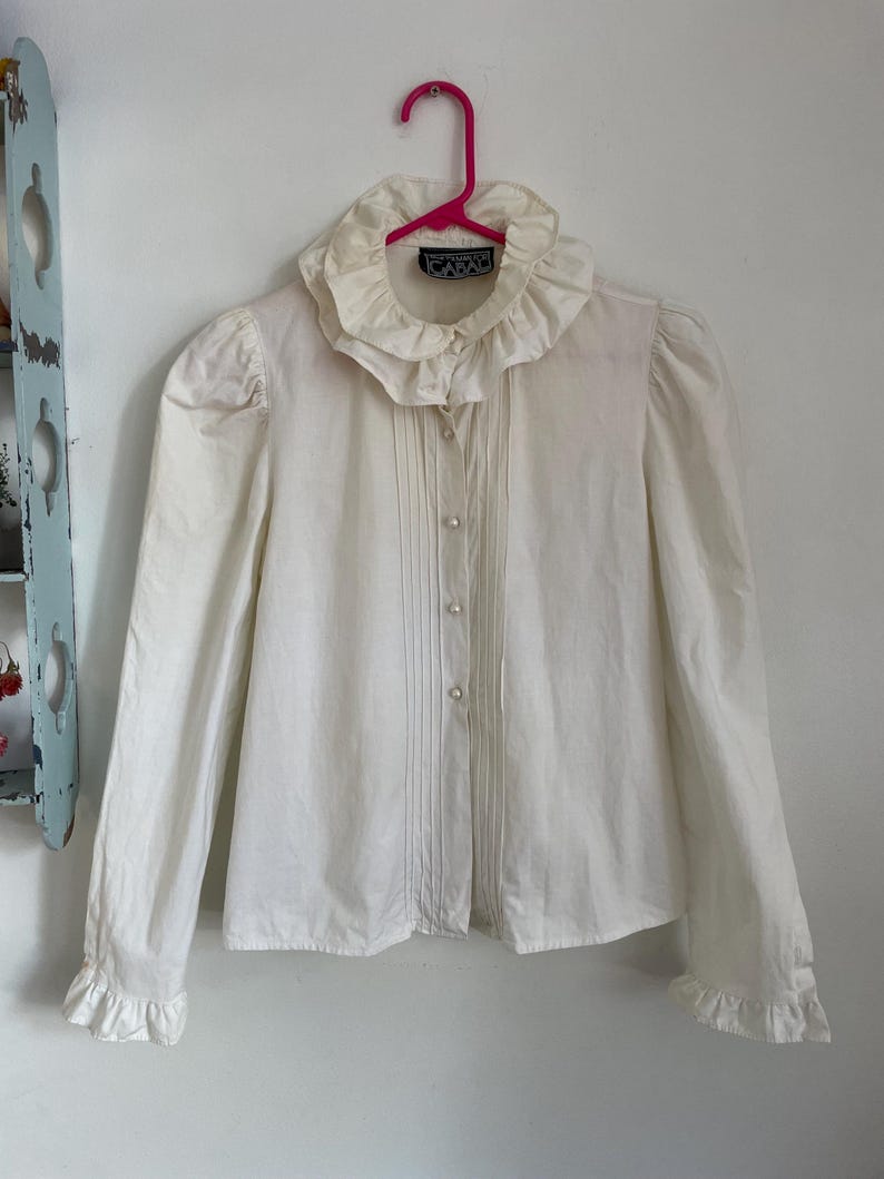 Puede incluir: Una blusa blanca de manga larga con cuello y pu&ntilde;os con volantes. La blusa tiene un cierre de botones en la parte delantera y est&aacute; plisada en la parte delantera. La etiqueta dice "CARAD"
