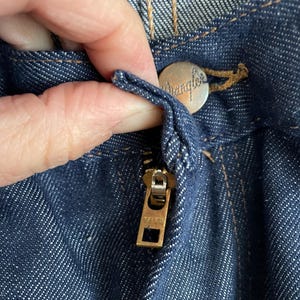 May include: Close-up of dark blue denim jeans with a brass zipper and a silver button. The button is embossed with the word "Wrangler". The zipper is marked "YLC". The stitching is a contrasting tan color.