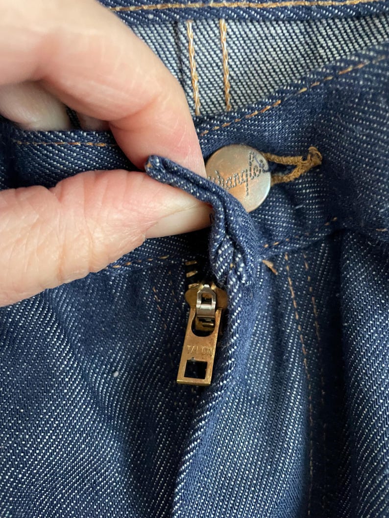 May include: Close-up of dark blue denim jeans with a brass zipper and a silver button. The button is embossed with the word "Wrangler". The zipper is marked "YLC". The stitching is a contrasting tan color.