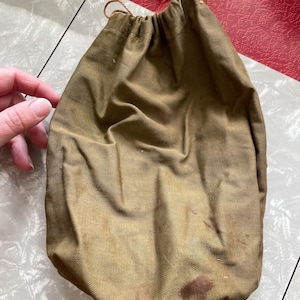 May include: A tan drawstring bag made of a textured fabric. The bag has a rounded bottom and a drawstring closure at the top. There are several dark stains on the bag. The bag is on a tiled surface.