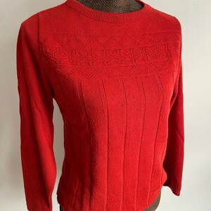 May include: A bright red sweater with a crew neck and long sleeves. The sweater features a textured design across the chest and vertical ribbing on the lower portion. The garment is displayed on a mannequin.