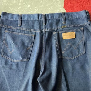 May include: Dark blue denim jeans with the Wrangler logo on a brown leather patch. The jeans have classic five-pocket styling, belt loops, and a worn appearance. The back pockets feature the signature Wrangler stitching.