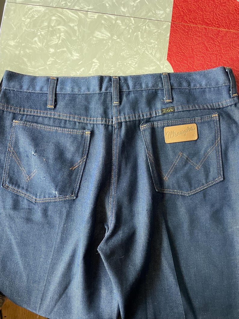 May include: Dark blue denim jeans with the Wrangler logo on a brown leather patch. The jeans have classic five-pocket styling, belt loops, and a worn appearance. The back pockets feature the signature Wrangler stitching.