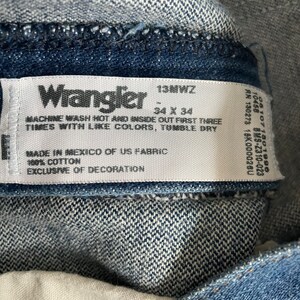 May include: A white care label with black text for Wrangler jeans. The label says "13MWZ", "34 x 34", "Machine wash hot and inside out first three times with like colours, tumble dry", "Made in Mexico of US fabric", "100% cotton", and "Exclusive of decoration".