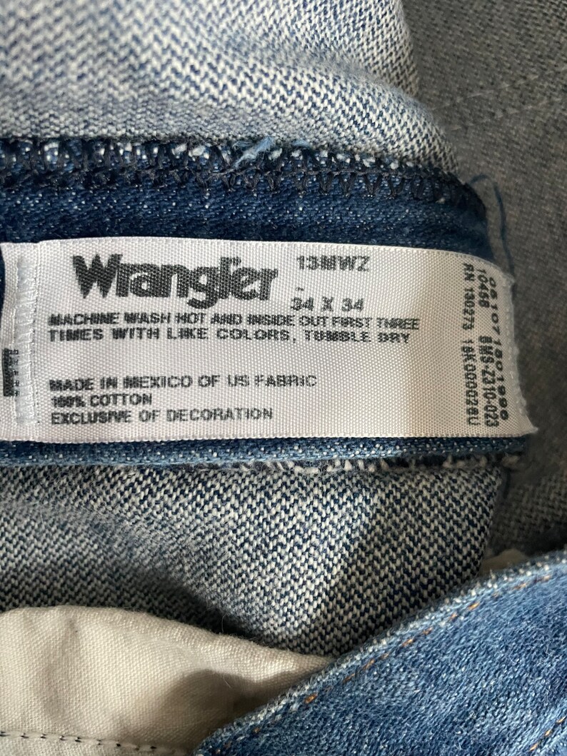 May include: A white care label with black text for Wrangler jeans. The label says "13MWZ", "34 x 34", "Machine wash hot and inside out first three times with like colours, tumble dry", "Made in Mexico of US fabric", "100% cotton", and "Exclusive of decoration".