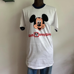 Op de afbeelding: Een wit ringer-t-shirt met donkerblauwe biesjes bij de kraag en mouwen. De voorkant toont een afbeelding van het gezicht van Mickey Mouse boven de rode tekst Walt Disney World, inclusief een klein wereldbolicoon in de letter D van Disney.