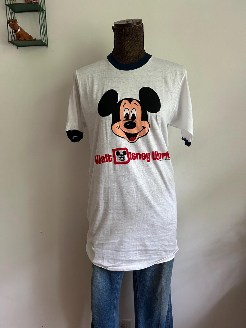 Op de afbeelding: Een wit ringer-t-shirt met donkerblauwe biesjes bij de kraag en mouwen. De voorkant toont een afbeelding van het gezicht van Mickey Mouse boven de rode tekst Walt Disney World, inclusief een klein wereldbolicoon in de letter D van Disney.