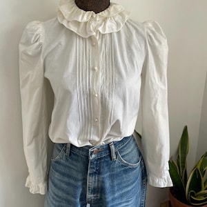 Puede incluir: Una blusa blanca de botones con cuello de volantes y mangas largas. La blusa tiene un frente plisado y est&aacute; hecha de una tela ligera.