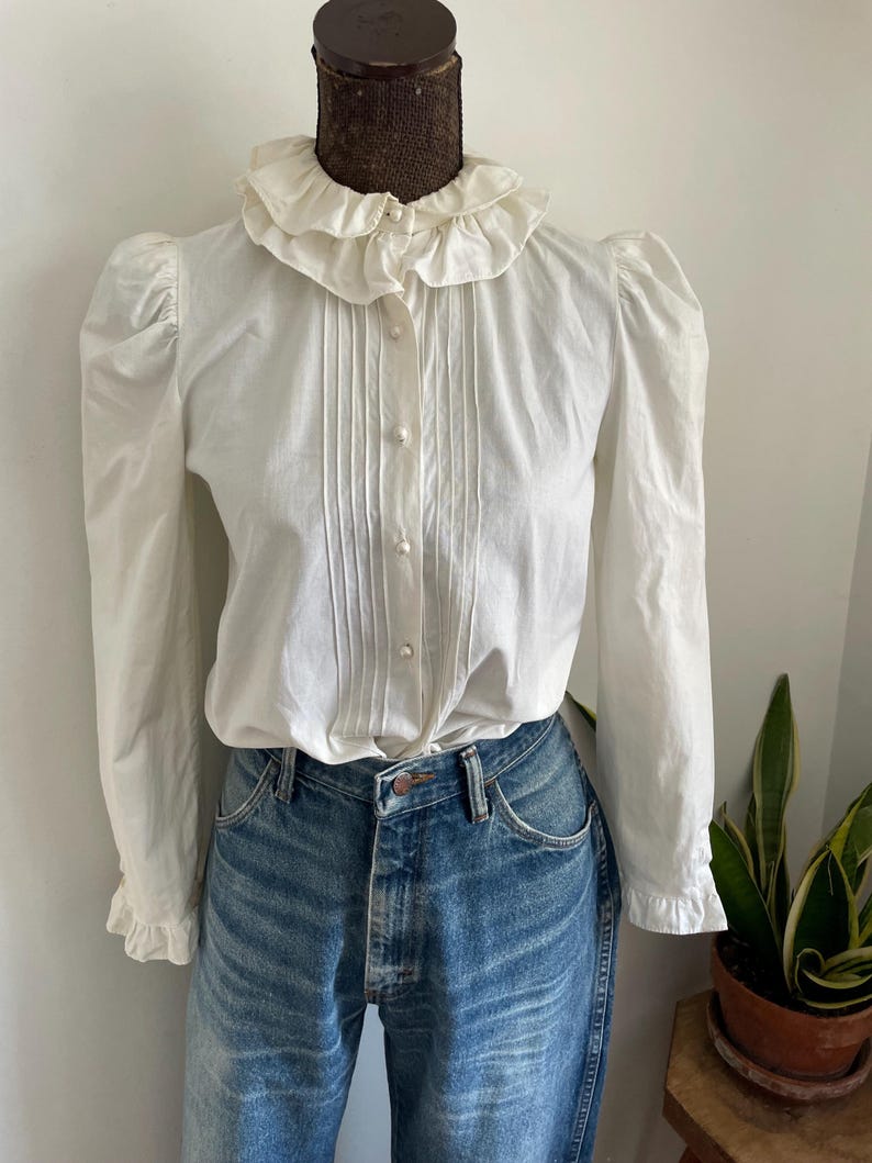 Puede incluir: Una blusa blanca de botones con cuello de volantes y mangas largas. La blusa tiene un frente plisado y est&aacute; hecha de una tela ligera.