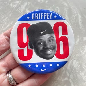 May include: A red, white, and blue button with a picture of a man wearing a black baseball cap with a white Nike swoosh. The button says "GRIFFEY" and "96" in large red letters. There is a white Nike swoosh at the bottom of the button.