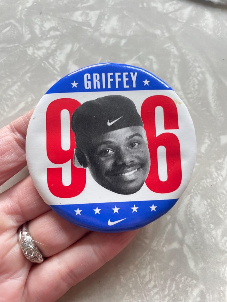 May include: A red, white, and blue button with a picture of a man wearing a black baseball cap with a white Nike swoosh. The button says "GRIFFEY" and "96" in large red letters. There is a white Nike swoosh at the bottom of the button.