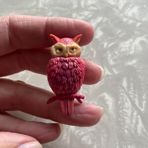 May include: A pink owl brooch with yellow eyes and a detailed feathered design. The owl is perched on a branch with its wings spread.