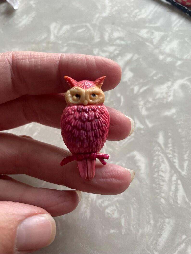 May include: A pink owl brooch with yellow eyes and a detailed feathered design. The owl is perched on a branch with its wings spread.