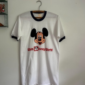 Op de afbeelding: Een wit ringer-t-shirt met donkerblauwe biesjes bij de kraag en mouwen. De voorkant toont een afbeelding van het gezicht van Mickey Mouse boven de rode tekst "Walt Disney World", waarbij een Mickey-icoon in een rood vierkant de letter D vervangt.