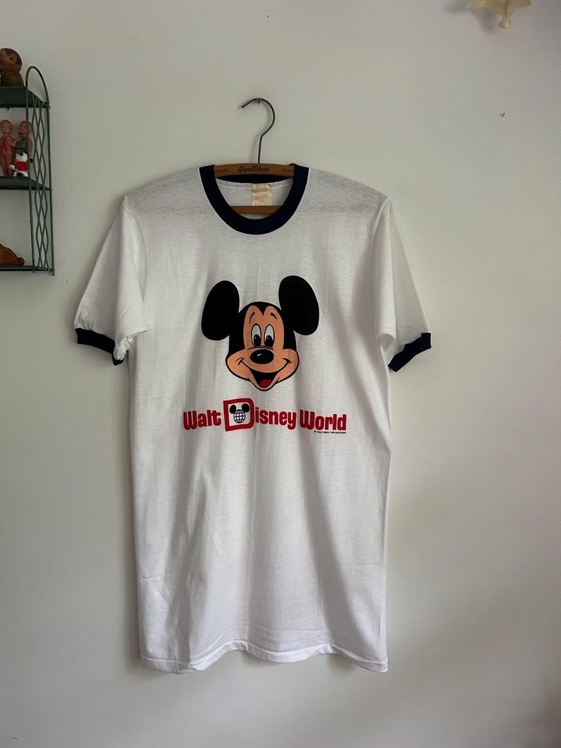 Op de afbeelding: Een wit ringer-t-shirt met donkerblauwe biesjes bij de kraag en mouwen. De voorkant toont een afbeelding van het gezicht van Mickey Mouse boven de rode tekst "Walt Disney World", waarbij een Mickey-icoon in een rood vierkant de letter D vervangt.