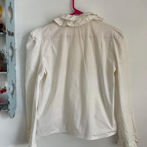Puede incluir: Una blusa blanca de manga larga con cuello de volantes y mangas abullonadas. La blusa est&aacute; colgada en una percha rosa.