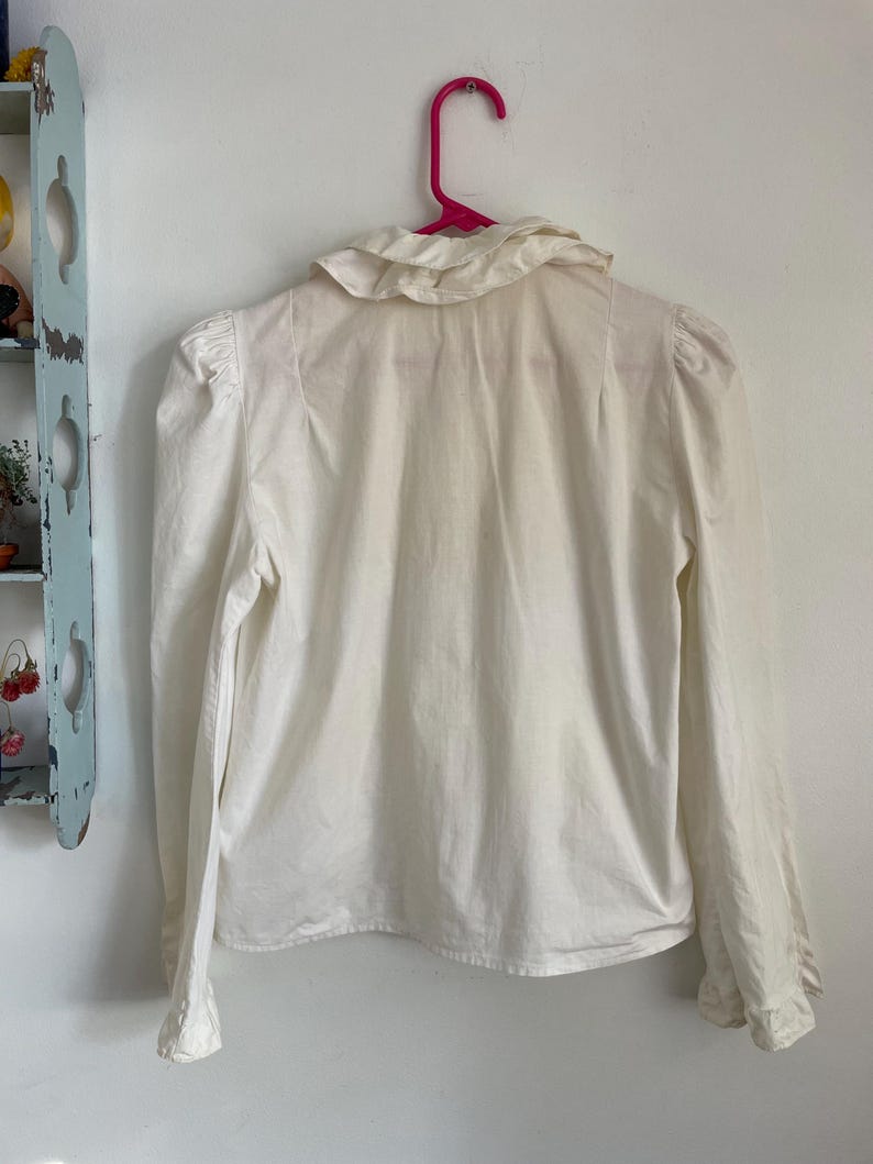 Puede incluir: Una blusa blanca de manga larga con cuello de volantes y mangas abullonadas. La blusa est&aacute; colgada en una percha rosa.