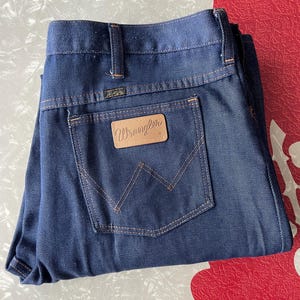 May include: Dark blue denim jeans with the Wrangler logo on the back pocket. The jeans have a classic five-pocket design with a straight leg. The stitching is a contrasting tan color. The jeans are folded neatly.