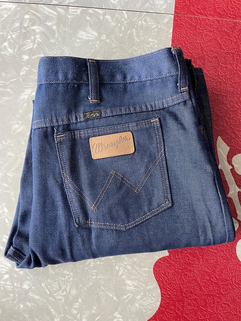 May include: Dark blue denim jeans with the Wrangler logo on the back pocket. The jeans have a classic five-pocket design with a straight leg. The stitching is a contrasting tan color. The jeans are folded neatly.