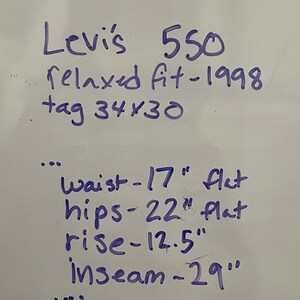 May include: Handwritten notes on a white surface, detailing the measurements of a pair of Levi's 550 relaxed fit jeans. The measurements include waist, hips, rise, and inseam in inches. The tag size is 34x30 and the jeans are from 1998.