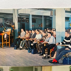 May include: A classroom scene with a teacher in a navy blue uniform standing at a desk with a microphone. Students are seated in rows of desks, listening to the teacher. The classroom has a light blue and white color scheme.